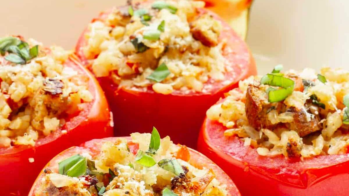 Close-up of stuffed tomatoes filled with rice, herbs, and browned toppings, arranged on a plate with a spoon. The tomatoes are garnished with fresh green basil leaves.