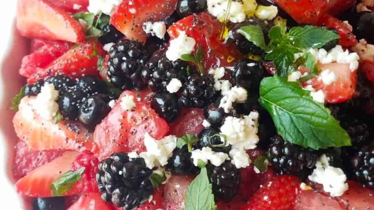 A vibrant salad featuring sliced strawberries, blackberries, crumbled cheese, and fresh mint leaves. The ingredients are drizzled with olive oil and seasoned with cracked black pepper.