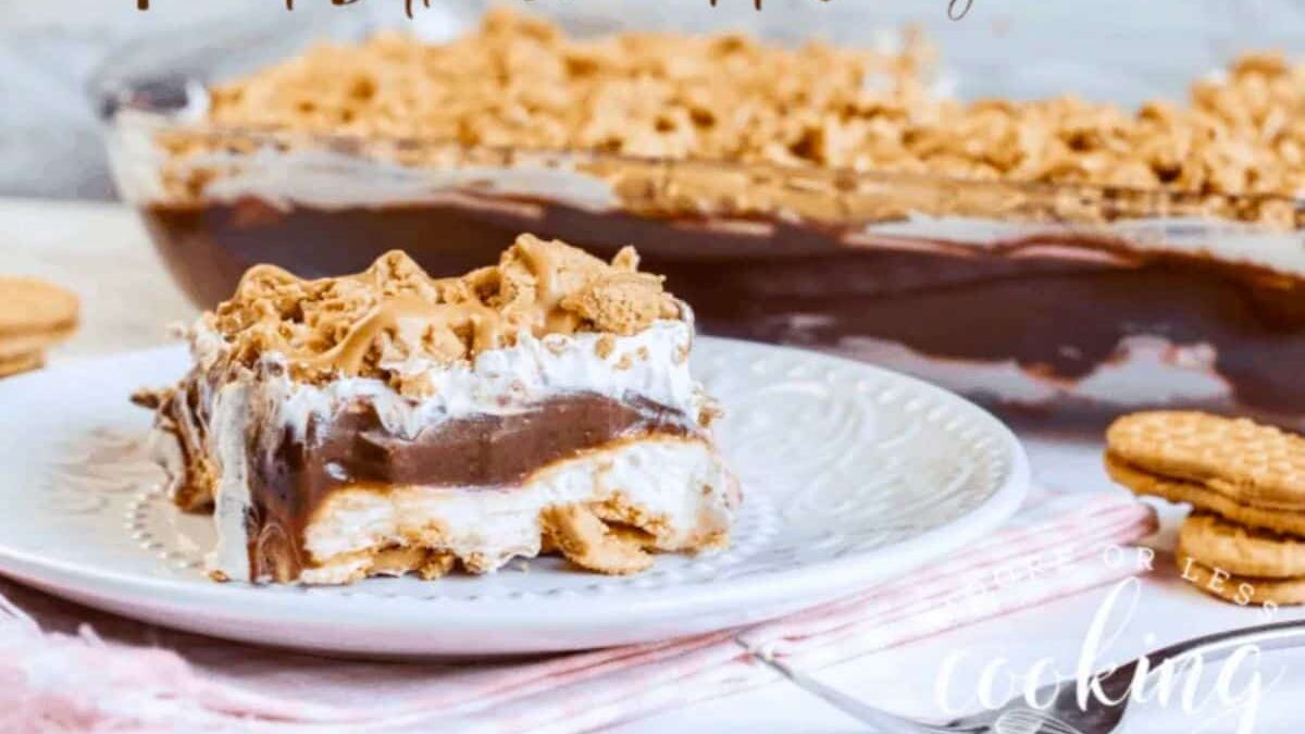 A slice of peanut butter chocolate lasagna on a white plate, featuring layers of creamy chocolate and white filling, topped with crumbled peanut butter cookies. The full dish is in the background with more cookies on the table.