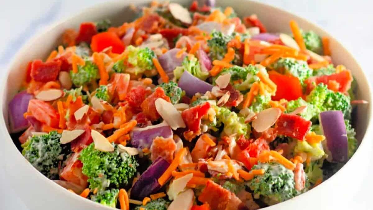A vibrant salad in a white bowl featuring broccoli, red onion slices, diced red bell peppers, shredded cheddar cheese, crispy bacon bits, and almond slivers. The colorful ingredients create a fresh and appetizing appearance.