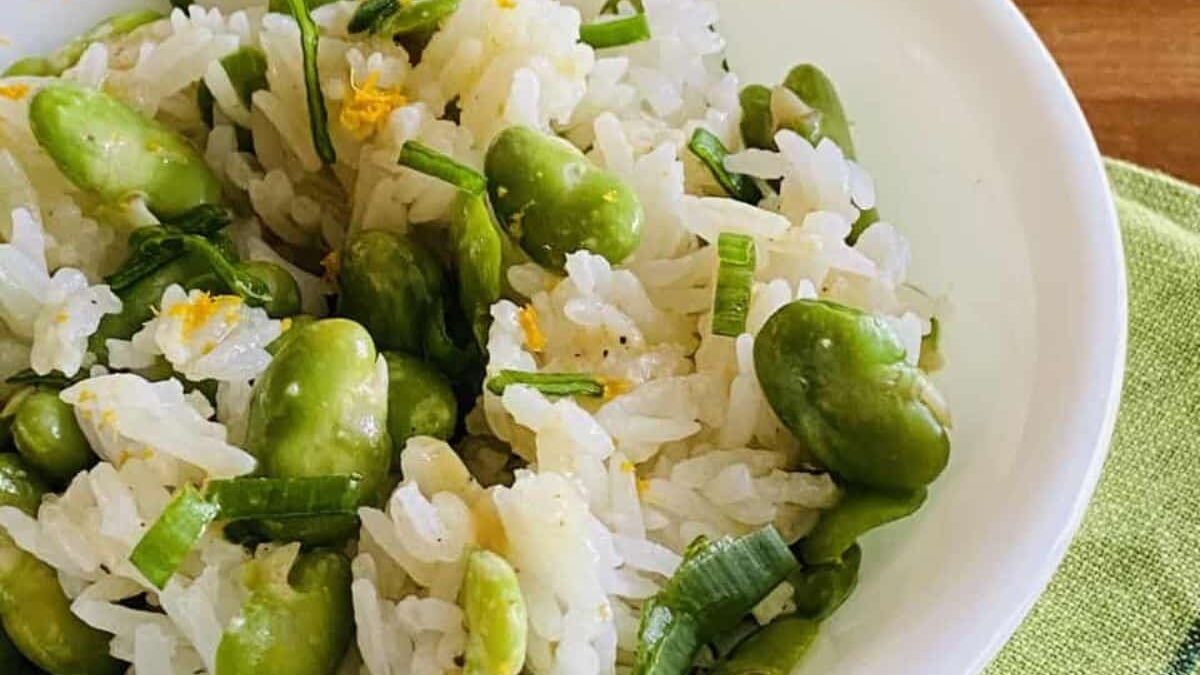 A bowl of rice mixed with broad beans and garnished with chopped green onions. The dish is presented on a white plate, placed on a green cloth. The rice appears fluffy, and the beans add a pop of green color.
