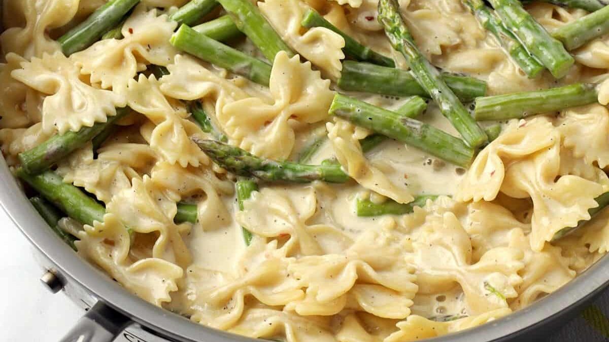 Close-up of creamy pasta with bow tie noodles and asparagus in a pan. The sauce appears rich and smooth, coating the pasta and bright green asparagus spears, offering a delicious and inviting presentation.