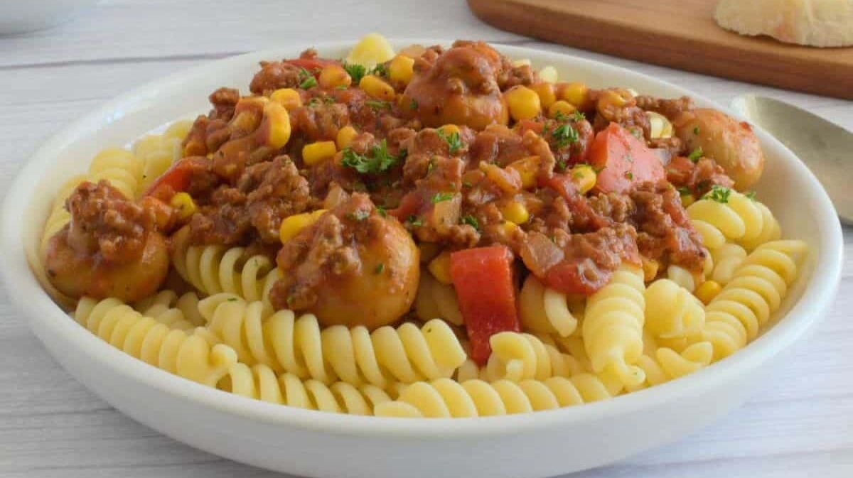 A white plate filled with rotini pasta topped with a tomato-based sauce containing ground beef, corn, red peppers, and mushrooms, garnished with herbs. In the background, there's a wooden board with slices of bread.