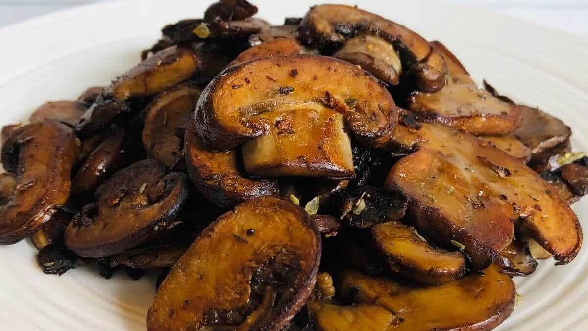A close-up of a white plate filled with sautéed sliced mushrooms, seasoned with herbs. The mushrooms are browned and glistening, creating a rustic and appetizing appearance.