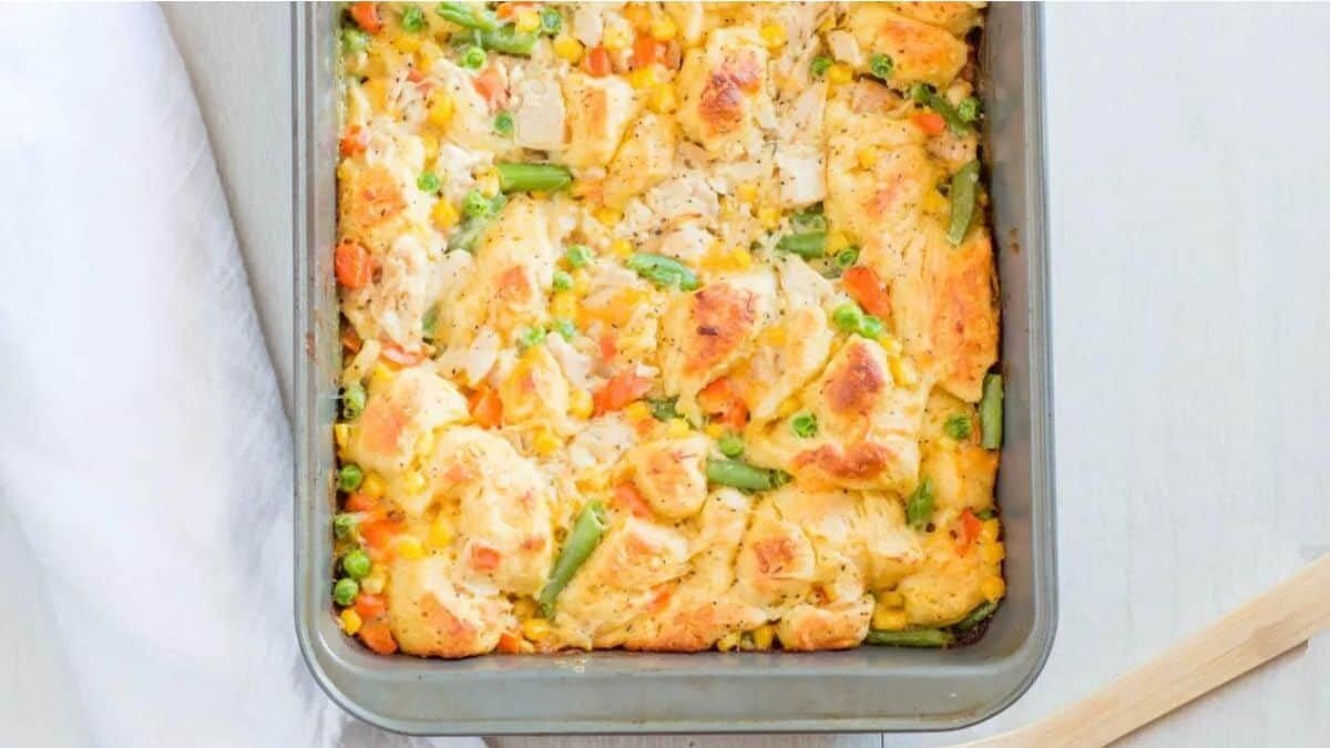 A casserole dish filled with a baked mixture of golden biscuit pieces, peas, carrots, green beans, corn, and a creamy sauce, sitting on a white surface next to a white napkin.
