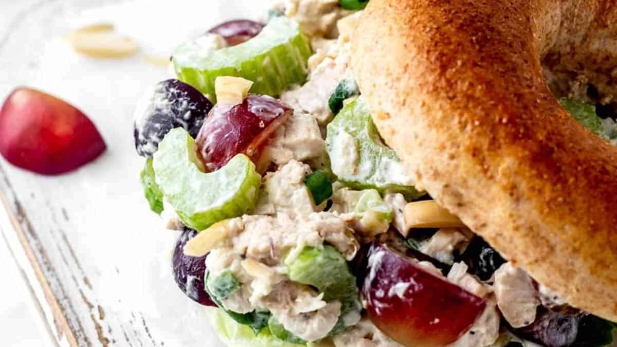 Close-up of a sandwich with a whole-grain bagel filled with a creamy chicken salad mixture. The salad includes grape halves, celery slices, and almond slivers on a lettuce leaf. The sandwich rests on a distressed white wooden surface.