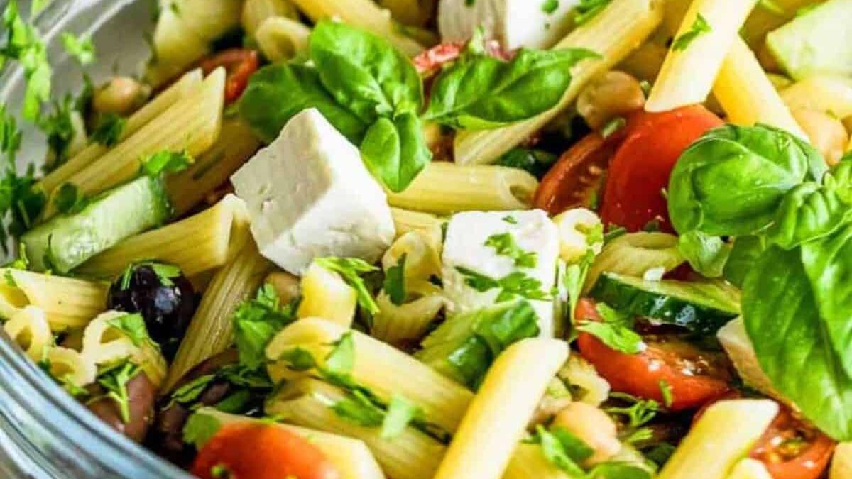 A colorful pasta salad with penne, cherry tomatoes, black olives, feta cheese cubes, cucumber slices, and fresh basil leaves garnished with chopped parsley in a clear bowl.