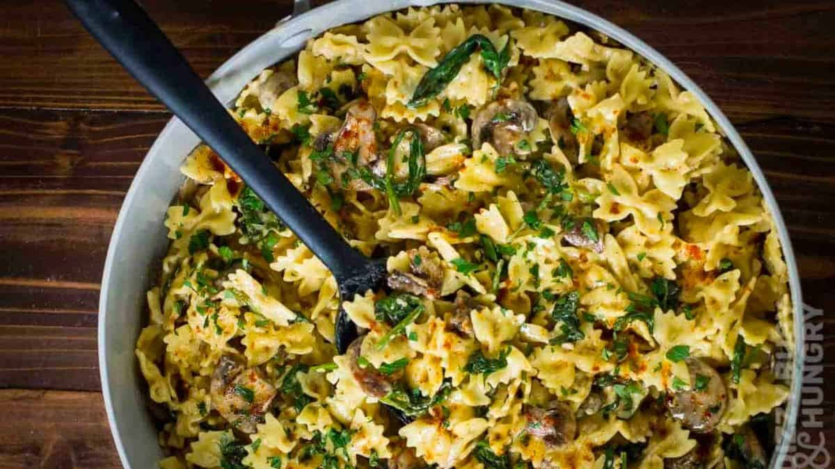 A dish of bowtie pasta mixed with sliced mushrooms, creamy sauce, and garnished with fresh herbs is presented in a white bowl. A serving spoon rests inside, ready to serve. The table surface is wooden.