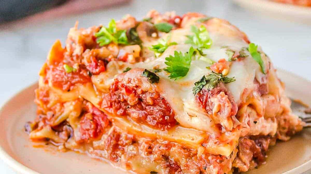 A close-up of a slice of lasagna on a beige plate. The layers include pasta, red tomato sauce, ground meat, and melted cheese, garnished with fresh cilantro. The lasagna is rich and juicy, with visible chunks of tomatoes and herbs.