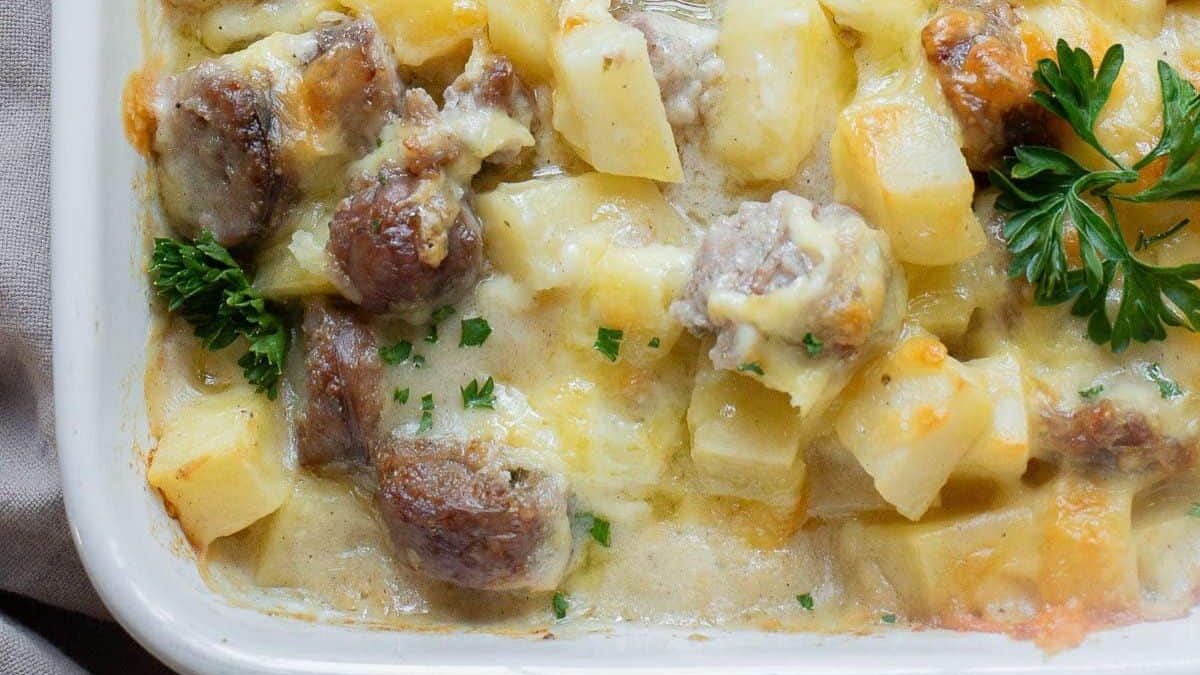 Close-up of a baked casserole dish filled with diced potatoes, browned sausage pieces, and a creamy cheese sauce. It's garnished with fresh parsley.
