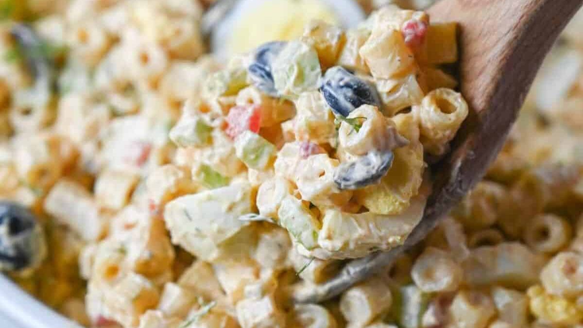 A wooden spoon scoops up creamy macaroni salad. The salad is filled with small pasta, sliced black olives, diced red peppers, and chopped celery, all mixed in a creamy dressing.