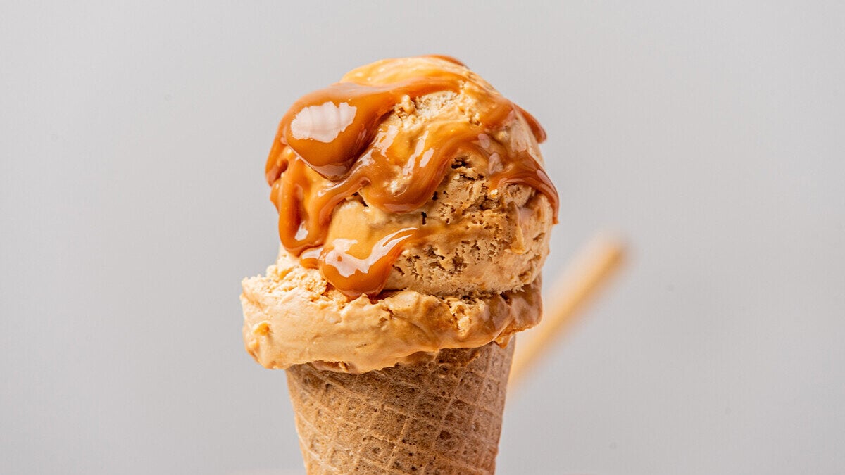A close-up of a scoop of caramel ice cream in a waffle cone. The ice cream is drizzled with a thick, glossy caramel sauce, creating a mouth-watering and indulgent treat against a plain, light gray background.