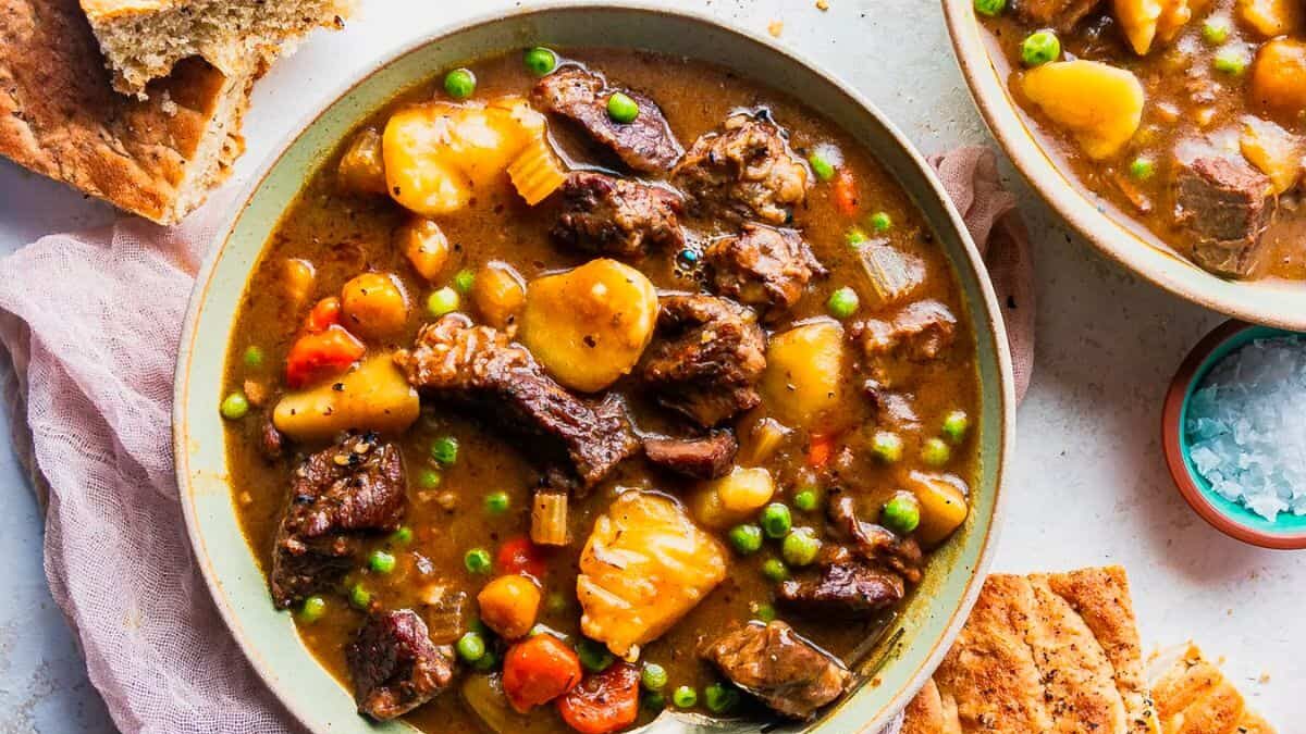 A bowl of hearty beef stew with chunks of beef, potatoes, carrots, peas, and celery in a rich broth. A spoon rests in the bowl, and slices of bread are on the side. A small dish of salt is nearby on a light cloth.