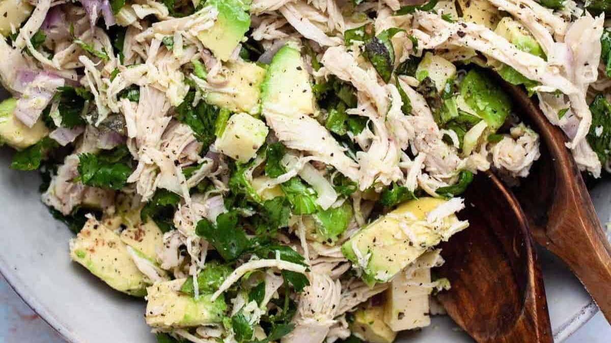 Close-up of a creamy chicken avocado salad with shredded chicken, avocado chunks, chopped cilantro, and red onions. The mixture is seasoned and served in a bowl with wooden utensils.
