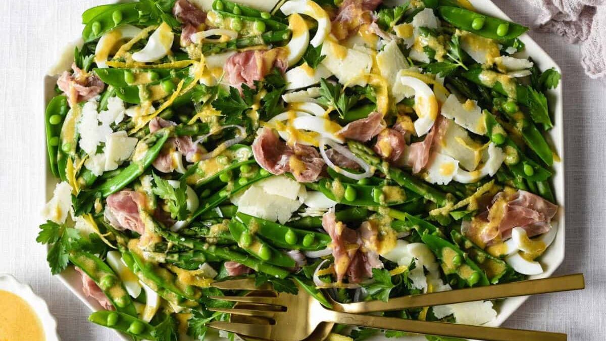 A plate of salad featuring sugar snap peas, sliced hard-boiled eggs, prosciutto, shaved Parmesan, and parsley, drizzled with a mustard vinaigrette. Two gold forks rest beside the colorful and fresh dish.