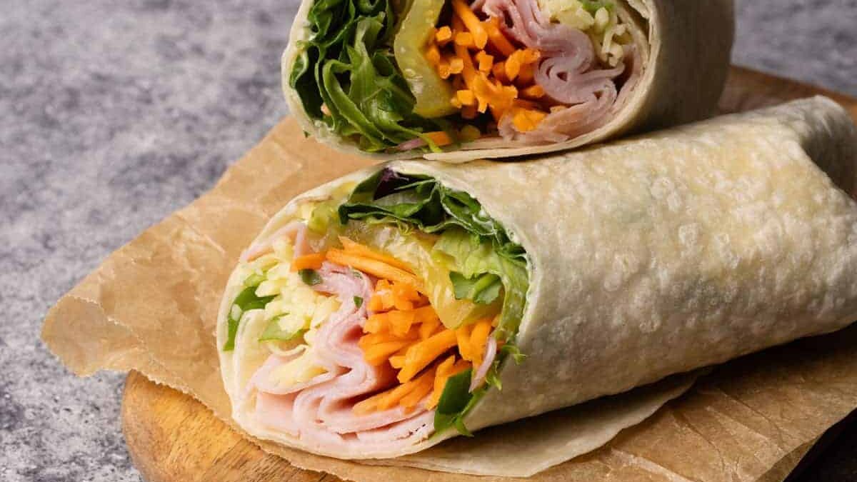 A close-up of a sliced wrap on a wooden board. The wrap is filled with sliced turkey, shredded cheese, lettuce, carrots, and other vegetables. It's placed on a piece of brown parchment paper.