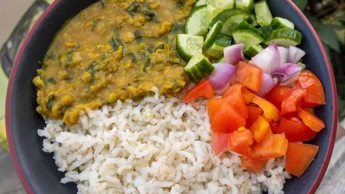 A bowl of white rice served with yellow lentil dal, chopped cucumber, diced red onion, and pieces of tomato, arranged in separate sections.