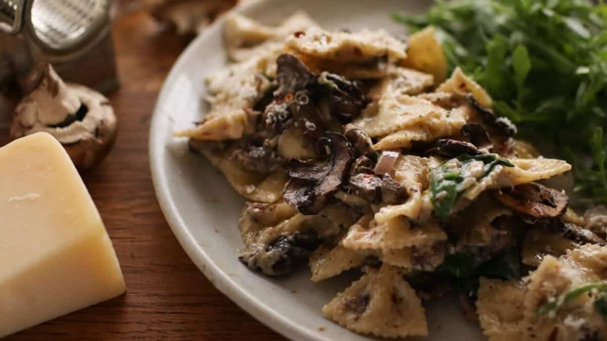 A plate of bowtie pasta with creamy mushroom sauce is garnished with fresh greens. Parmesan cheese and mushrooms are in the background on a wooden table.