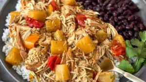 A plate filled with shredded chicken, pineapple chunks, and red bell pepper atop white rice, accompanied by a side of black beans. A fork rests on the plate, and a sprig of cilantro is visible nearby.