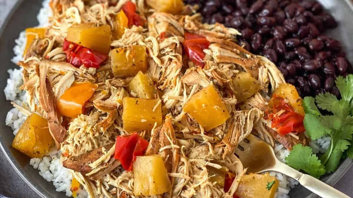 A plate filled with shredded chicken, pineapple chunks, and red bell pepper atop white rice, accompanied by a side of black beans. A fork rests on the plate, and a sprig of cilantro is visible nearby.