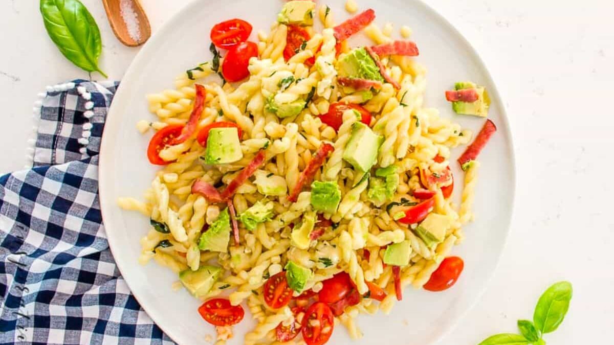 A colorful plate of pasta salad with spiral pasta, sliced cherry tomatoes, diced avocado, and small strips of salami. Garnished with fresh basil, served on a white plate with a checkered napkin on the side.