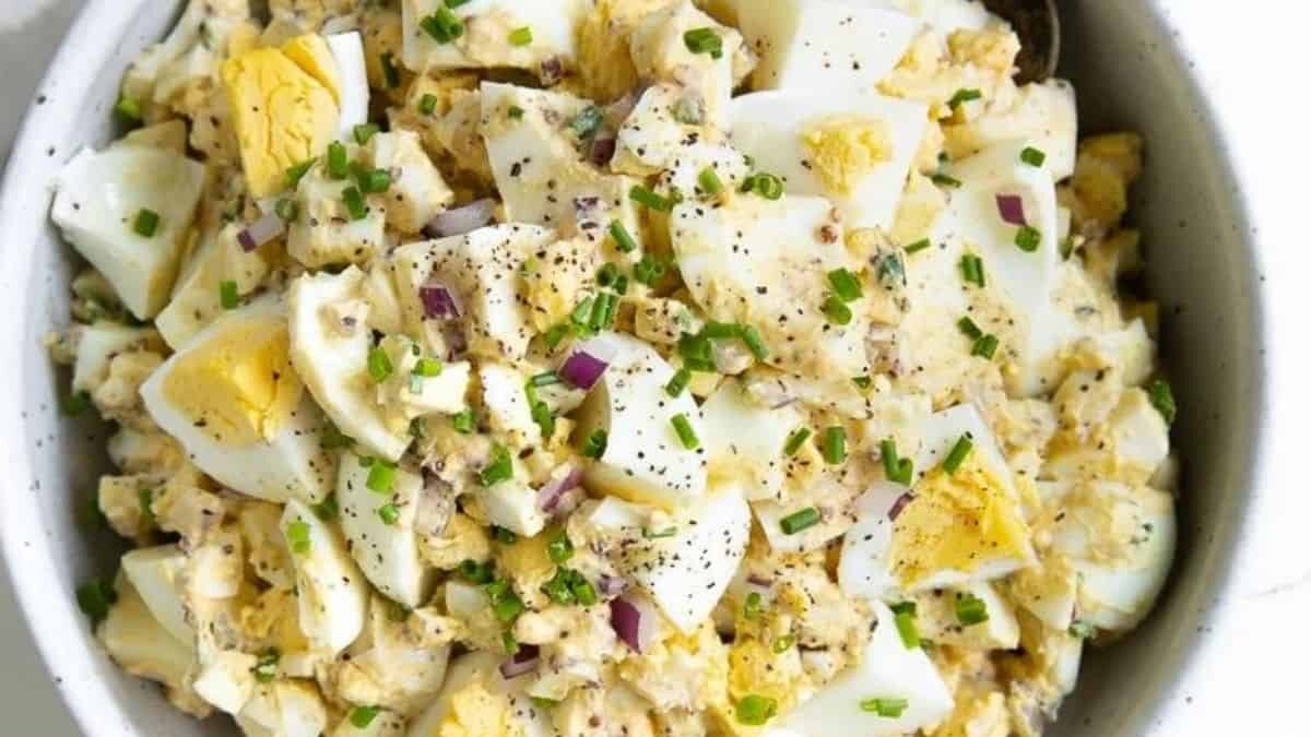 A bowl of egg salad with chopped boiled eggs, red onion, herbs, and black pepper. The creamy mixture is garnished with sliced chives, adding a pop of green to the dish.