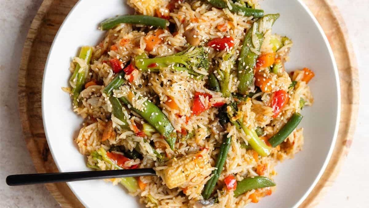 A white plate filled with vegetable stir-fried rice featuring green beans, broccoli, red bell peppers, and sprinkled with sesame seeds, with a black fork resting on the side.