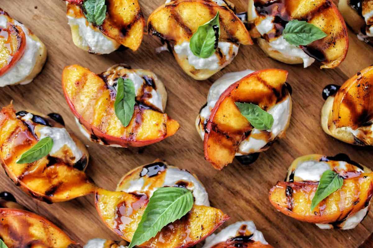 A wooden board topped with crostini, each layered with creamy cheese, grilled peach slices, fresh basil leaves, and drizzled with balsamic glaze.