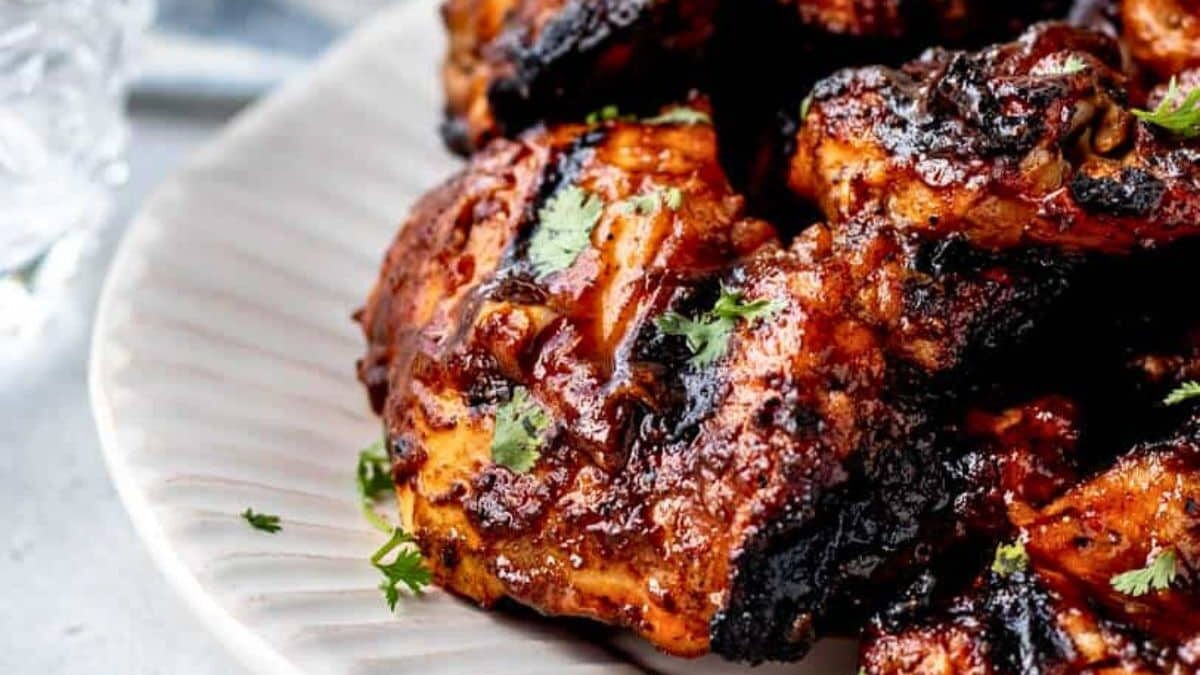 Close-up of grilled chicken pieces on a white plate. The chicken is charred and coated with a glossy BBQ sauce, garnished with fresh herbs, possibly cilantro. The background shows part of a glass and a blurred blue cloth.