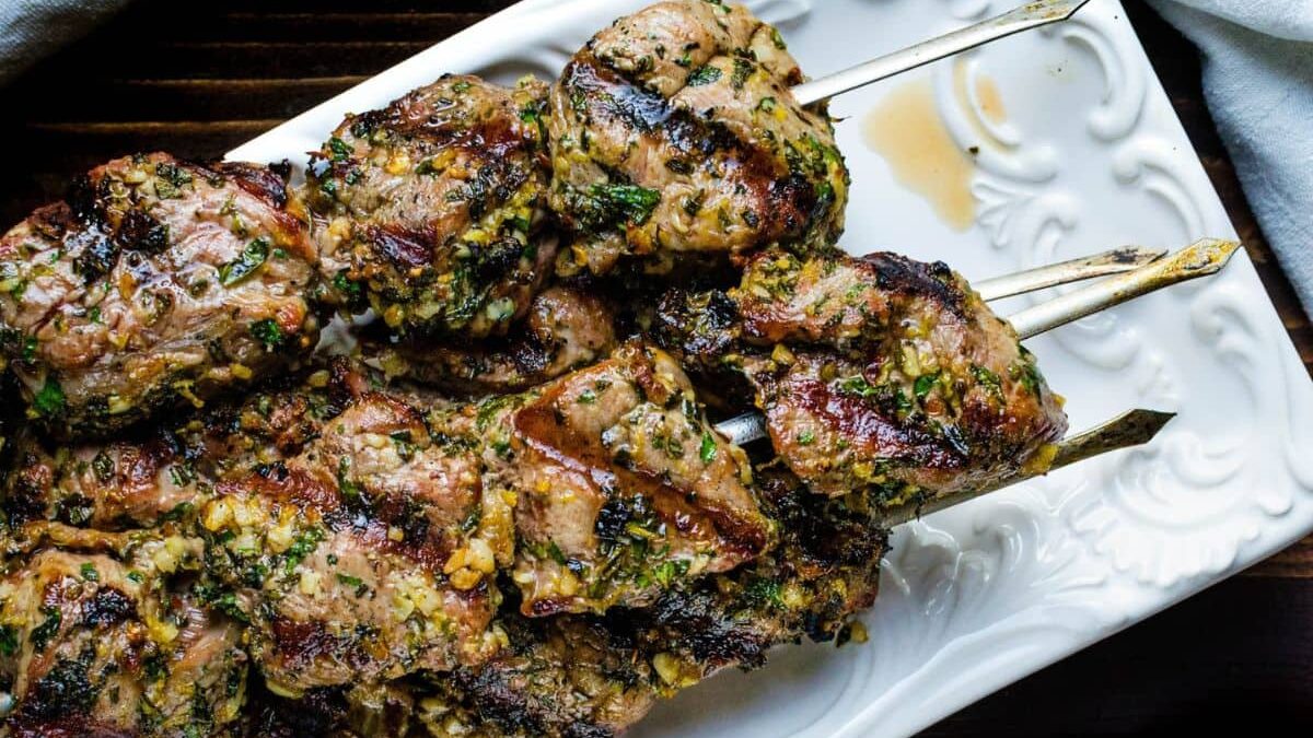 Grilled marinated meat skewers, seasoned with herbs and spices, served on a rectangular white platter. The meat appears juicy and charred, with visible pieces of garlic and parsley for added flavor.