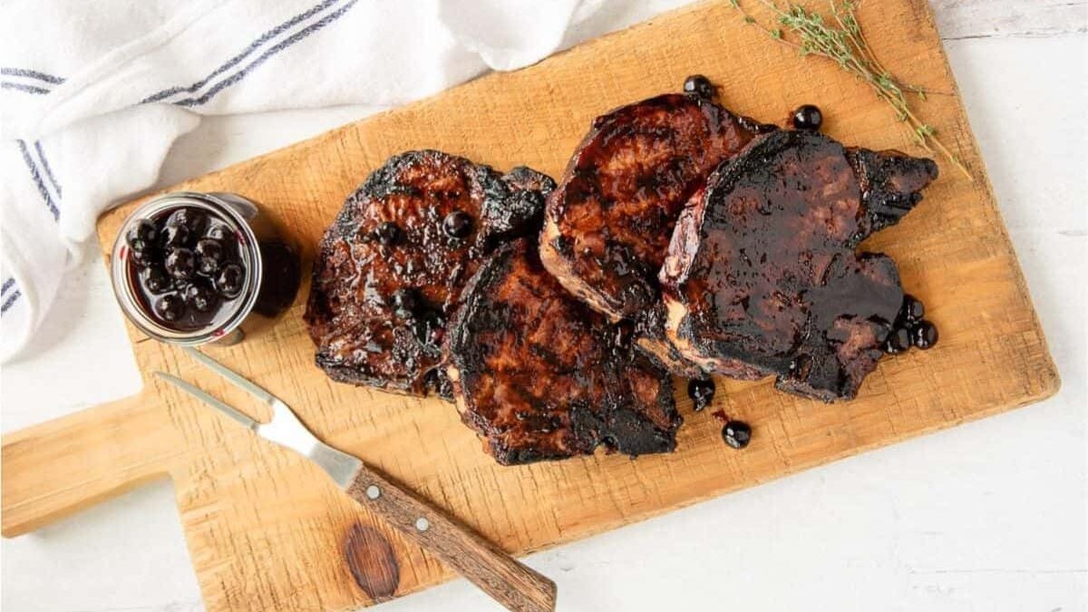 Grilled pork chops glazed with a dark sauce on a wooden cutting board, accompanied by a jar of black currant sauce and a small meat fork. A sprig of thyme rests beside the meat, and a white cloth with blue stripes is nearby.