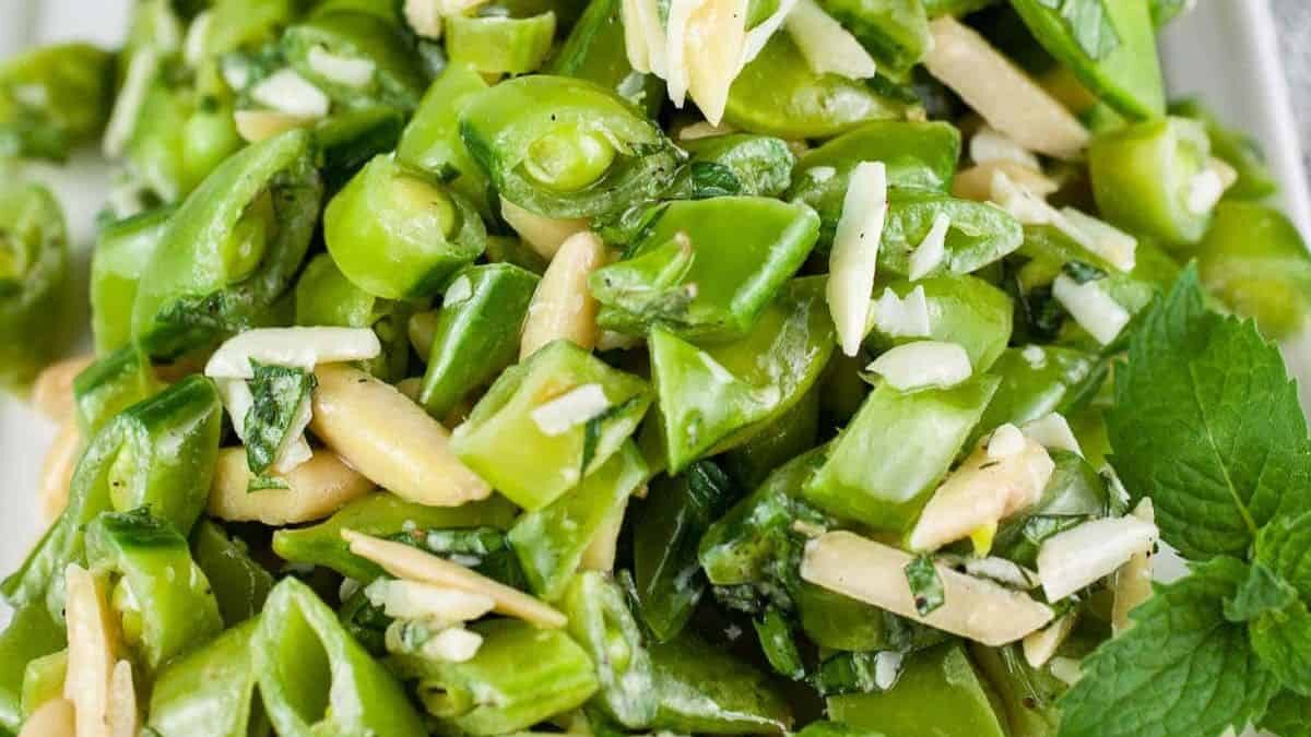 A close-up of a fresh green bean salad mixed with slivered almonds, chopped herbs, and a light dressing. The vibrant green color showcases the freshness of the beans, and there is a sprig of mint visible on the side.