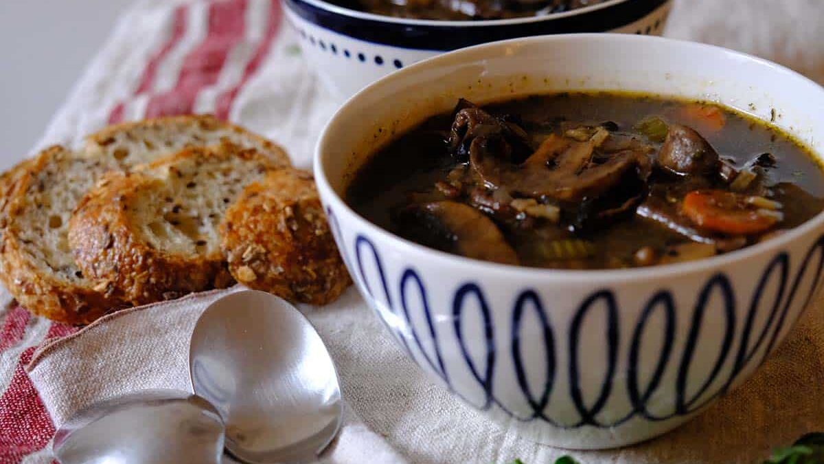 A bowl of mushroom and vegetable soup sits on a table alongside sliced multigrain bread. Two spoons lie nearby on a red and white cloth. Another bowl of soup can be seen in the background. Fresh herbs are also visible on the table.