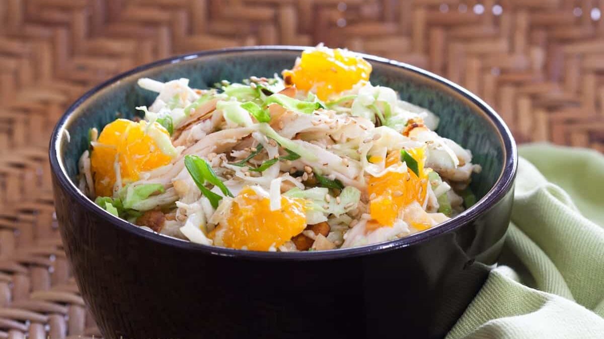 A vibrant salad in a black bowl features shredded cabbage, mandarin orange slices, sliced green onions, and sesame seeds. The bowl is set on a textured woven surface with a green cloth partially visible beside it.