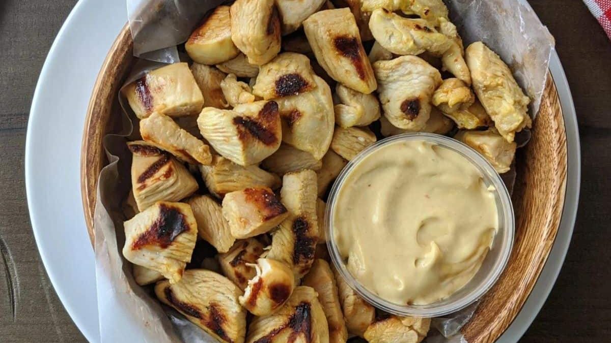 A wooden bowl filled with grilled chicken pieces, lined with parchment paper, placed on a white plate. A small glass bowl of creamy dipping sauce is on the side. The background is a wooden table with part of a red and white cloth.