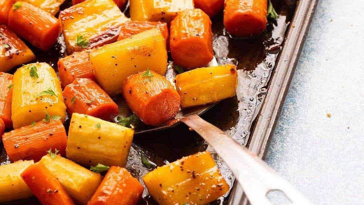 A baking tray filled with roasted carrots and parsnips, seasoned with herbs and drizzled with oil. A metal spatula is lifting some of the vegetables, revealing their caramelized edges. The background surface is a light blue.