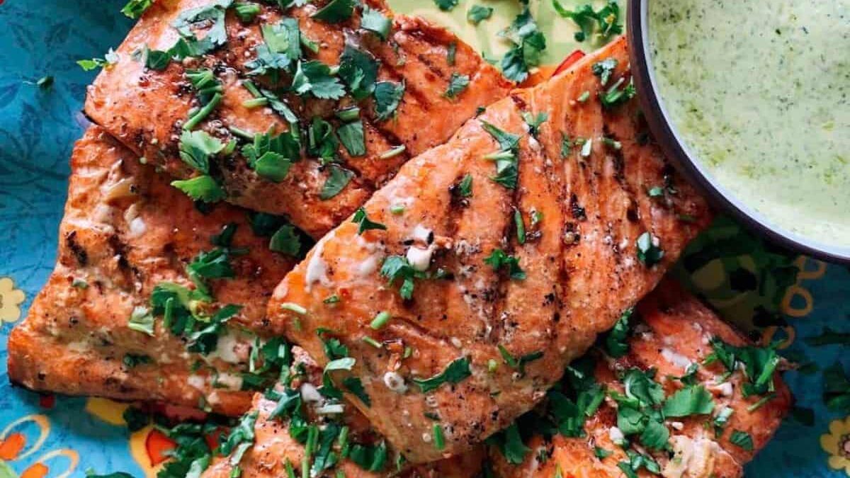 Grilled salmon fillets topped with chopped cilantro served on a colorful plate, accompanied by a bowl of creamy green sauce.