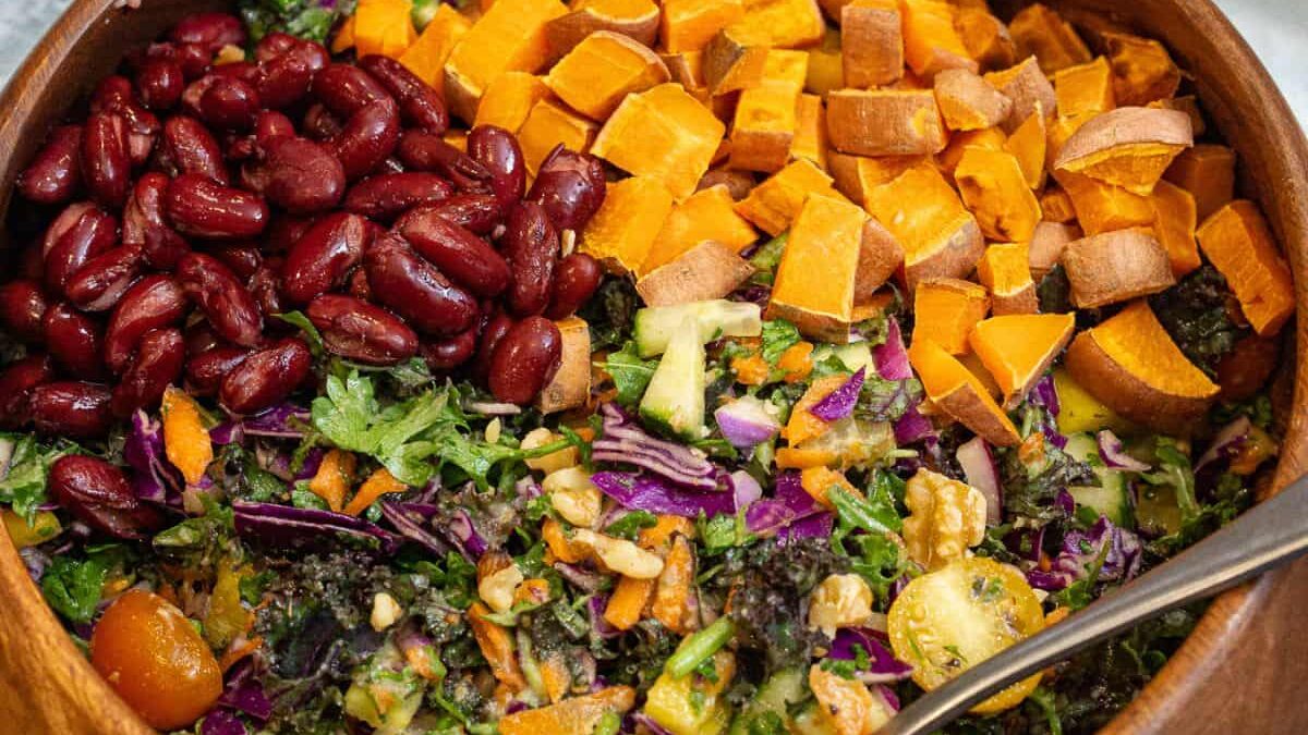 A wooden bowl filled with a colorful salad, including diced sweet potatoes, kidney beans, chopped kale, shredded carrots, purple cabbage, and sliced cucumbers. A spoon is placed on the side of the bowl.