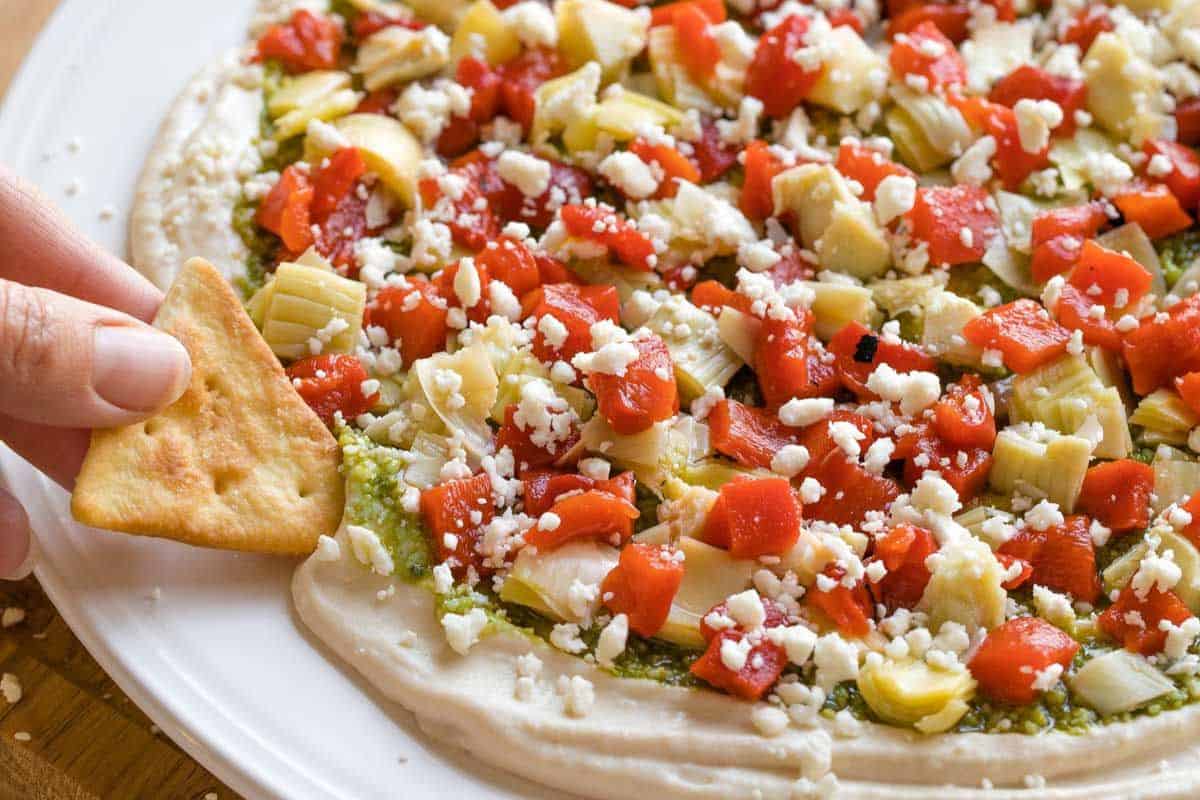 A hand dips a triangular cracker into a layered Mediterranean dip topped with diced red peppers, artichokes, crumbled feta cheese, and herbs on a white plate.