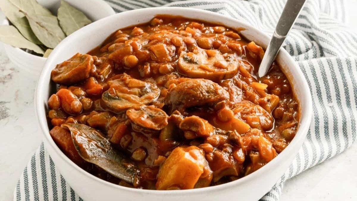 A bowl of hearty lentil stew with mushrooms and vegetables in a rich, savory sauce. The stew is set on a striped cloth, with bay leaves visible to the side, and a spoon resting in the bowl.