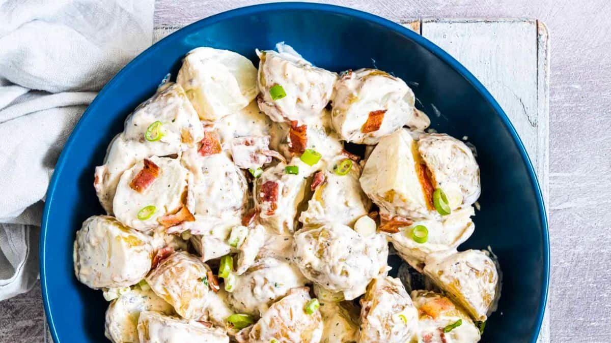 A blue bowl filled with creamy potato salad, garnished with chopped green onions and bacon bits. The salad is placed on a rustic white wooden surface with a light-colored cloth next to it.
