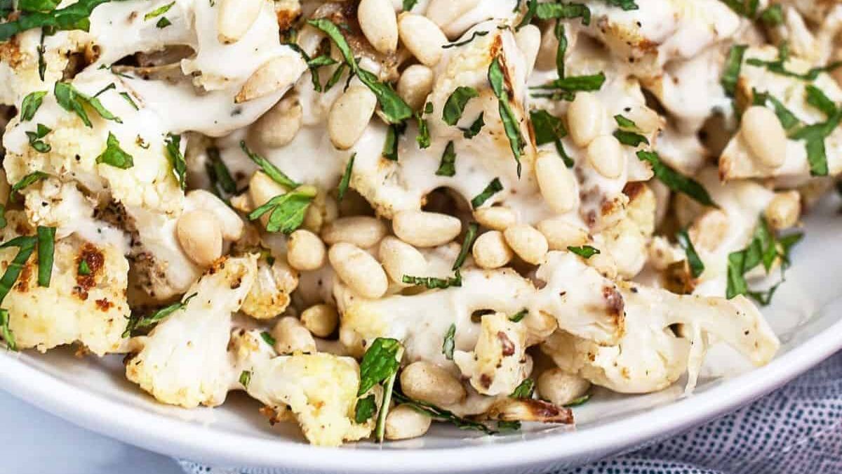 A close-up of roasted cauliflower florets topped with creamy tahini sauce, pine nuts, and chopped fresh herbs, served on a white plate.