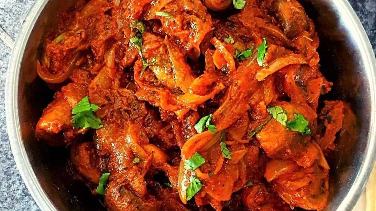 A stainless steel bowl filled with spicy and saucy chicken curry. The dish features tender chicken pieces coated in a rich, red sauce, garnished with fresh cilantro leaves. It's placed on a textured gray surface.