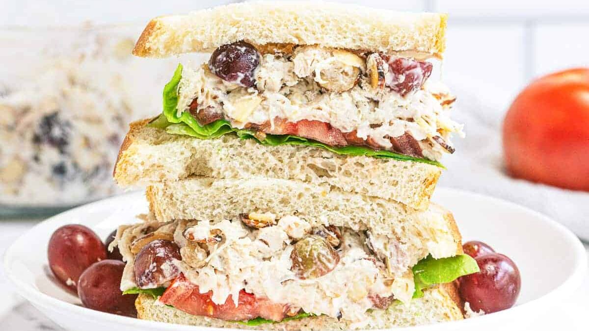 A sandwich stacked with three slices of bread, filled with chicken salad, lettuce, tomato slices, and grapes. The sandwich is placed on a white plate with additional grapes on the side.