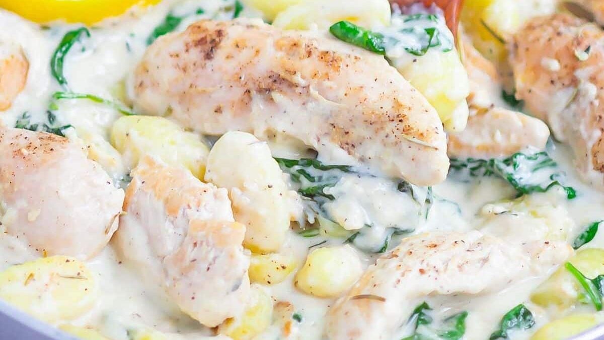 A creamy dish featuring cooked chicken pieces, gnocchi, and spinach in a white sauce. A slice of lemon is visible on the side, and a wooden spoon is partially submerged in the dish.