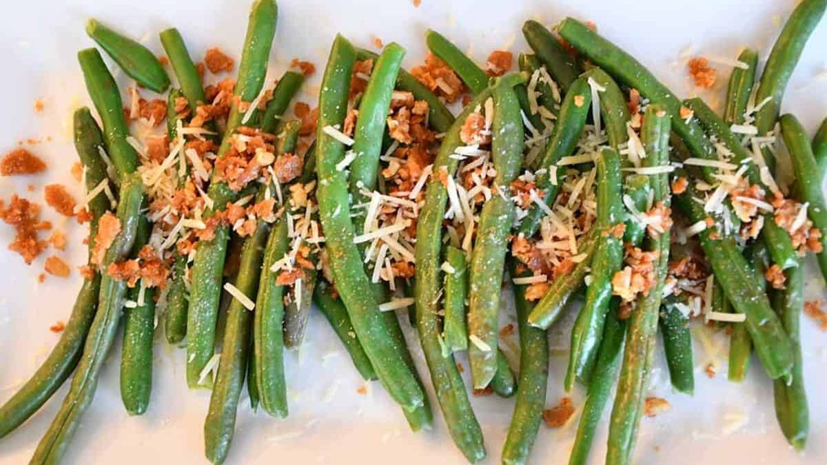 A dish of cooked green beans garnished with crumbled bacon and grated cheese, arranged on a white plate.