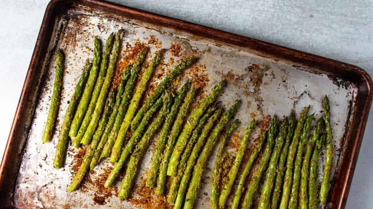 A baking sheet with roasted asparagus, lightly seasoned and arranged in rows. The asparagus tips are slightly browned, indicating they are tender and cooked. The baking sheet has a rustic, well-used appearance.