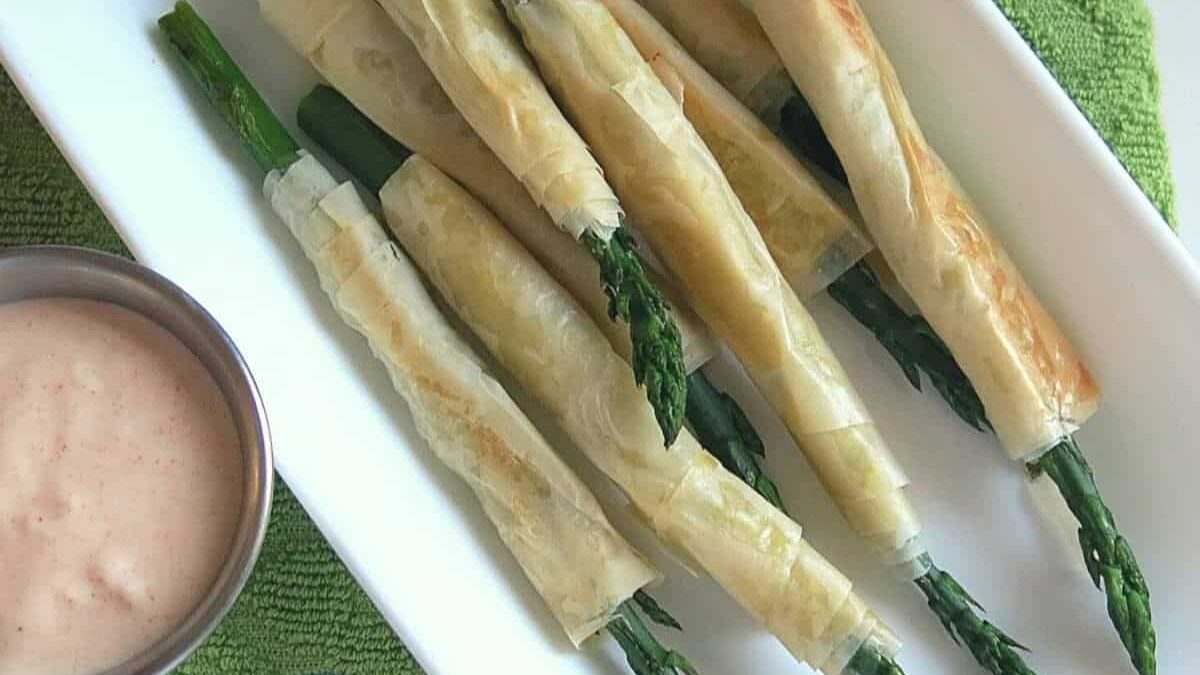 A white rectangular plate with phyllo-wrapped asparagus spears. The spears are golden brown and crispy. A small metal cup with pink dipping sauce is placed beside the plate. The background includes a green textured surface.