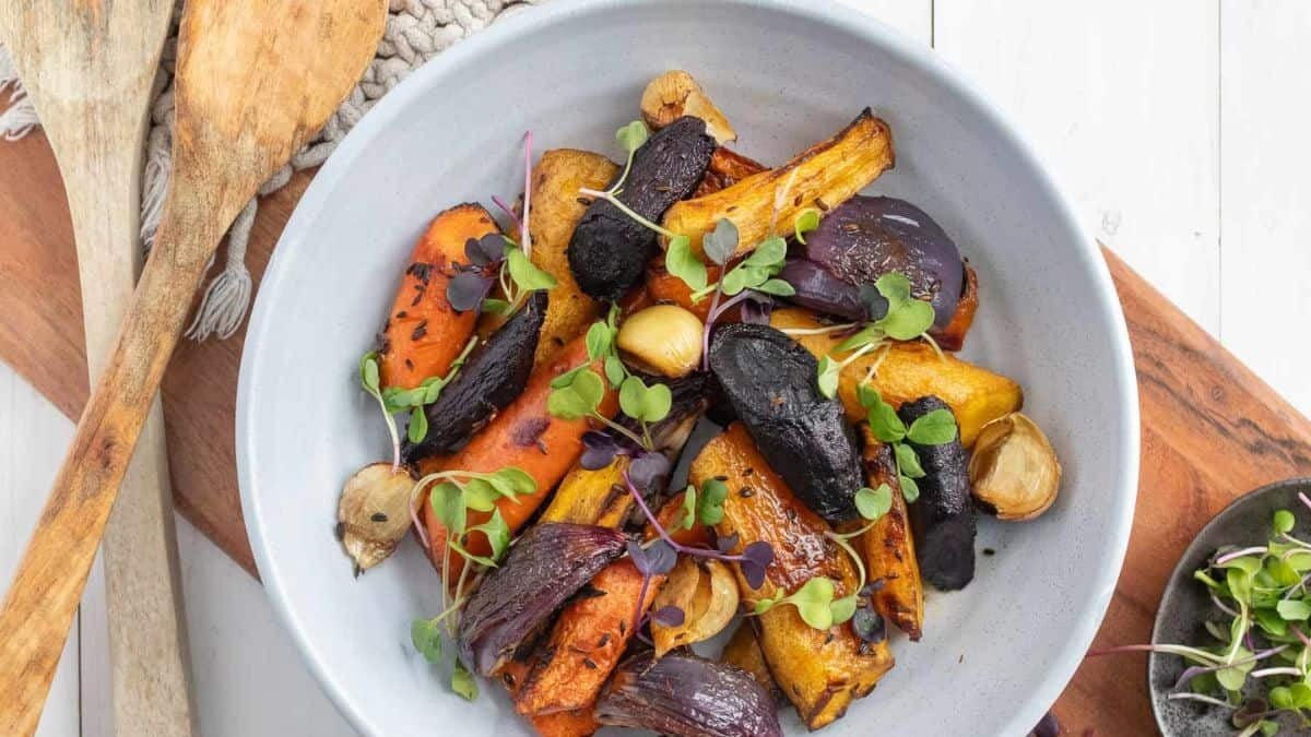 A white bowl filled with assorted roasted vegetables, including carrots, red onions, and possibly parsnips, garnished with fresh microgreens. The bowl is placed on a wooden board next to wooden utensils and a small dish of additional microgreens.