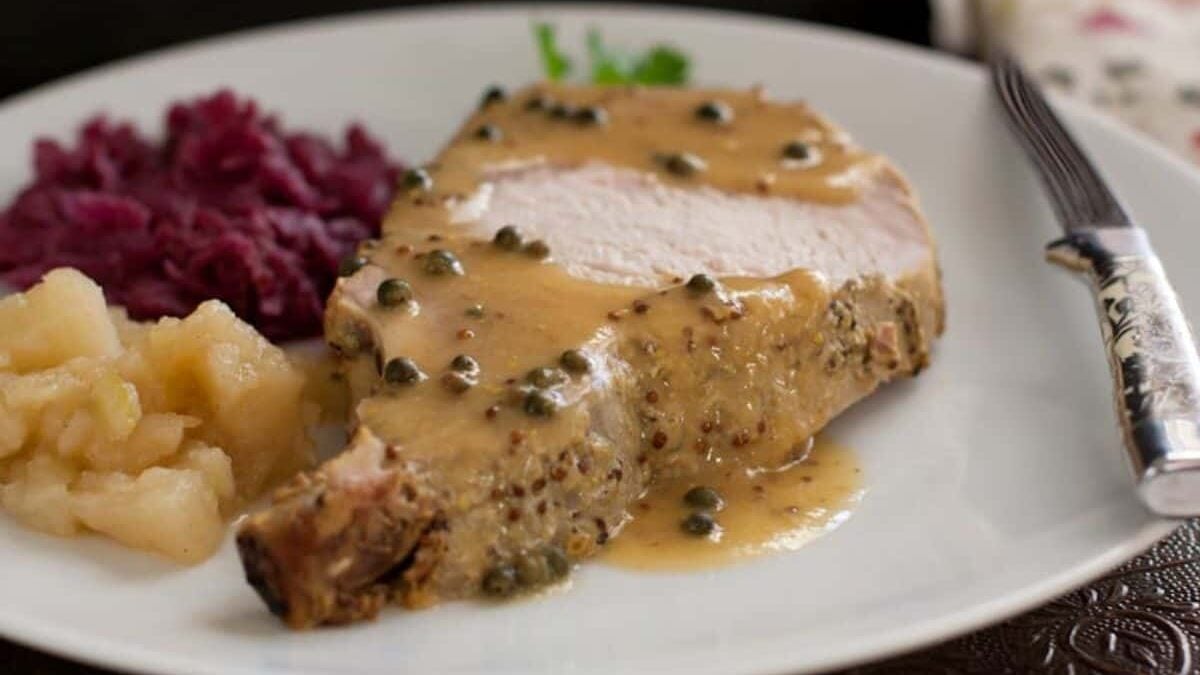 A plate with a slice of roasted pork topped with a creamy mustard caper sauce, served with sides of red cabbage and applesauce. A silver knife rests on the white plate next to the pork.